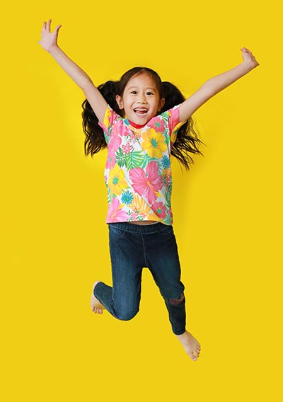 Happy Asian kindergarten girl, wearing a floral pattern summer shirt, jumping in excitement on a yellow background.